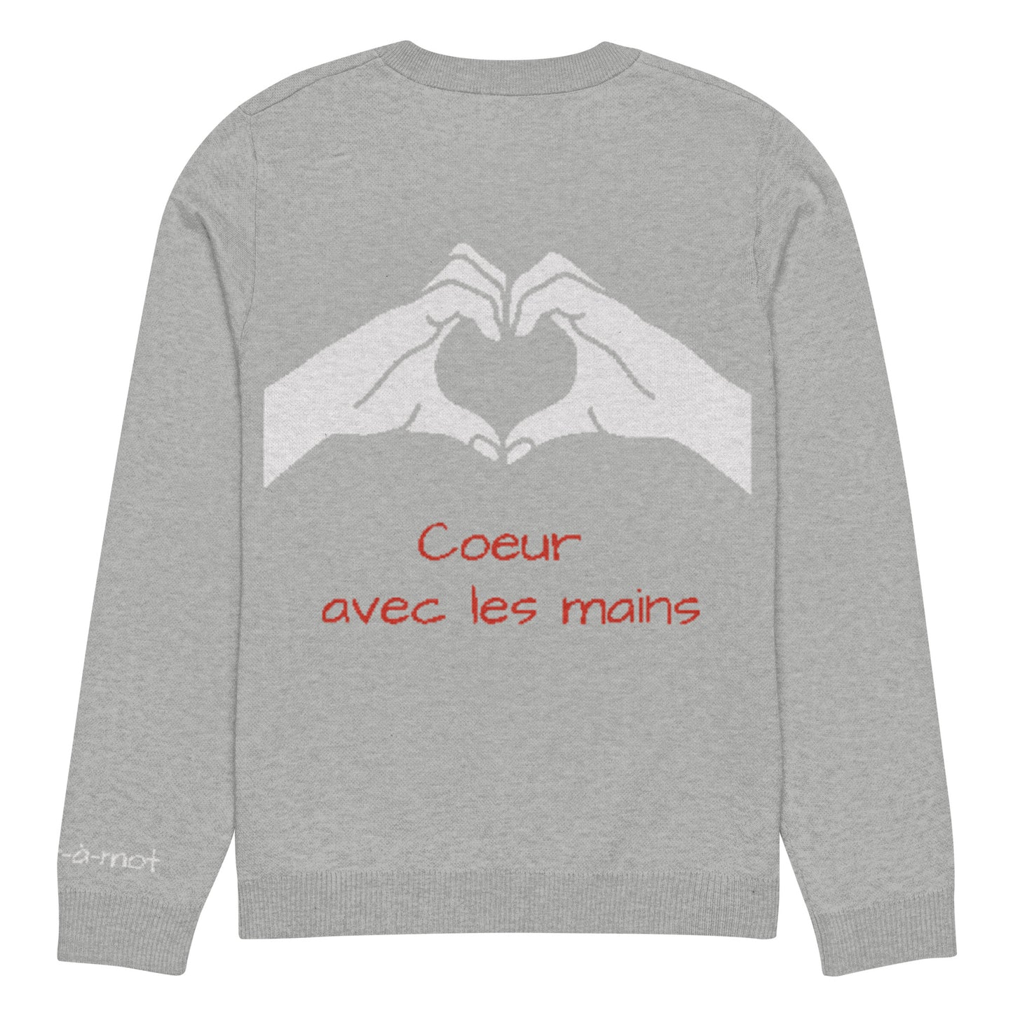 Back view of a light grey knitted jumper with a ribbed neckline, cuffs, and hem. Two white hands are shown gesturing a heart shape. Below the hand graphic, the phrase "Coeur avec les mains" is written in red. A small embroidered brand tag "mot-à-mot" is visible on the lower left sleeve