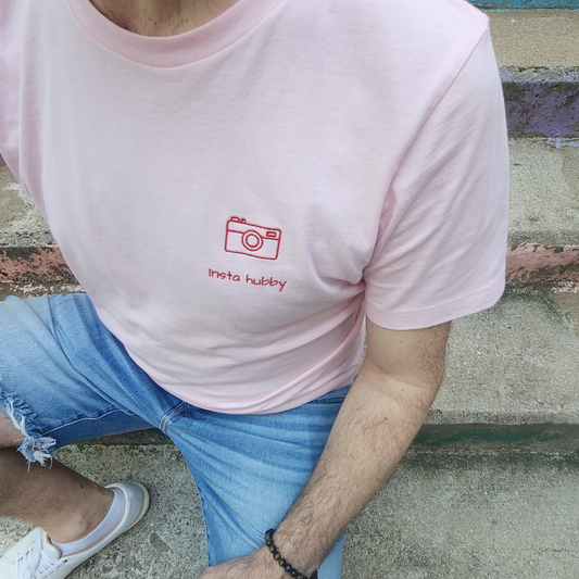 Front view of a light pink short-sleeved shirt featuring a small, red embroidered camera icon and the phrase "Insta Hubby" in a simple red font beneath it, located on the left side of the chest.