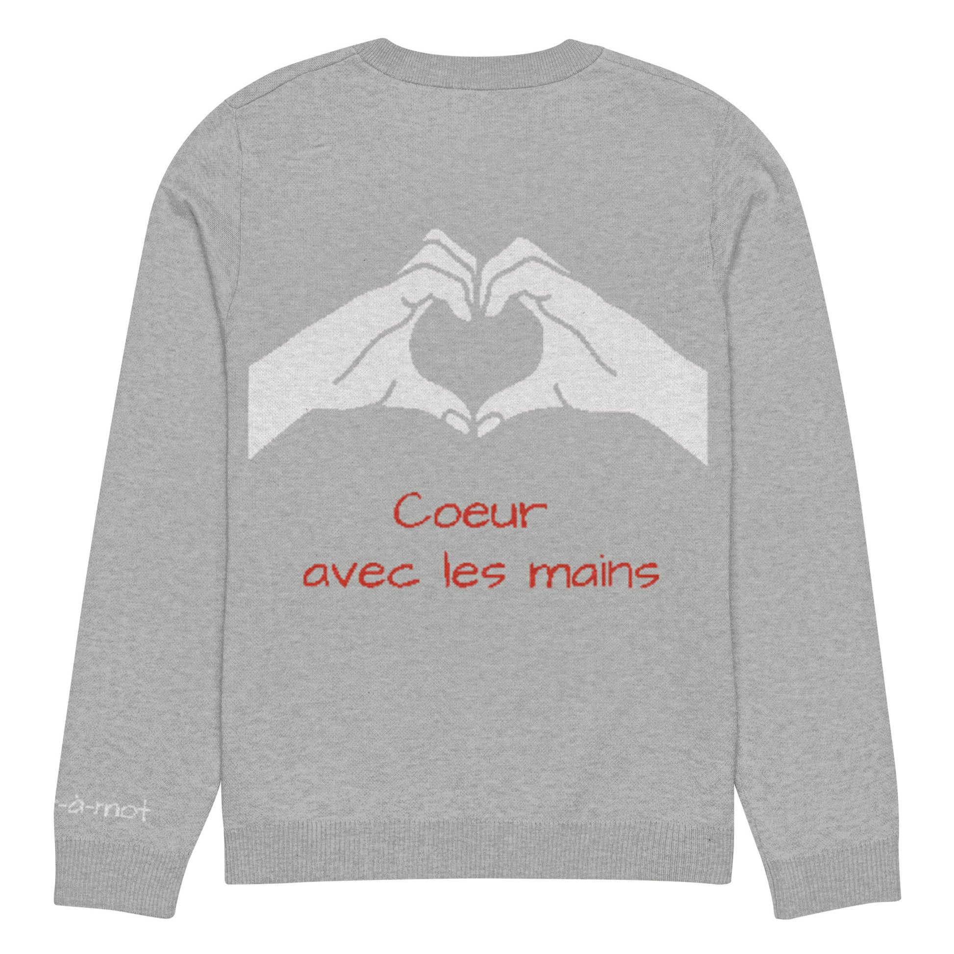 Back view of a light grey knitted jumper with a ribbed neckline, cuffs, and hem. Two white hands are shown gesturing a heart shape. Below the hand graphic, the phrase "Coeur avec les mains" is written in red. A small embroidered brand tag "mot-à-mot" is visible on the lower left sleeve
