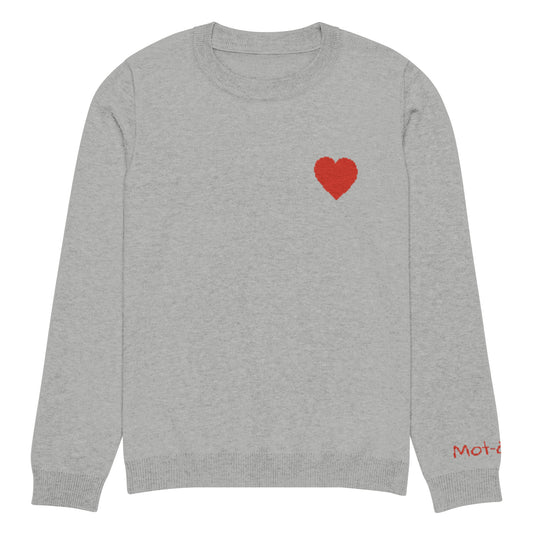 Front view of a grey knitted jumper with a ribbed neckline, cuffs, and hem. A prominent red heart design is centered on the chest, constructed from the knit itself (jacquard technique). A small red embroidered brand tag "mot-à-mot" is visible on the lower right sleeve.